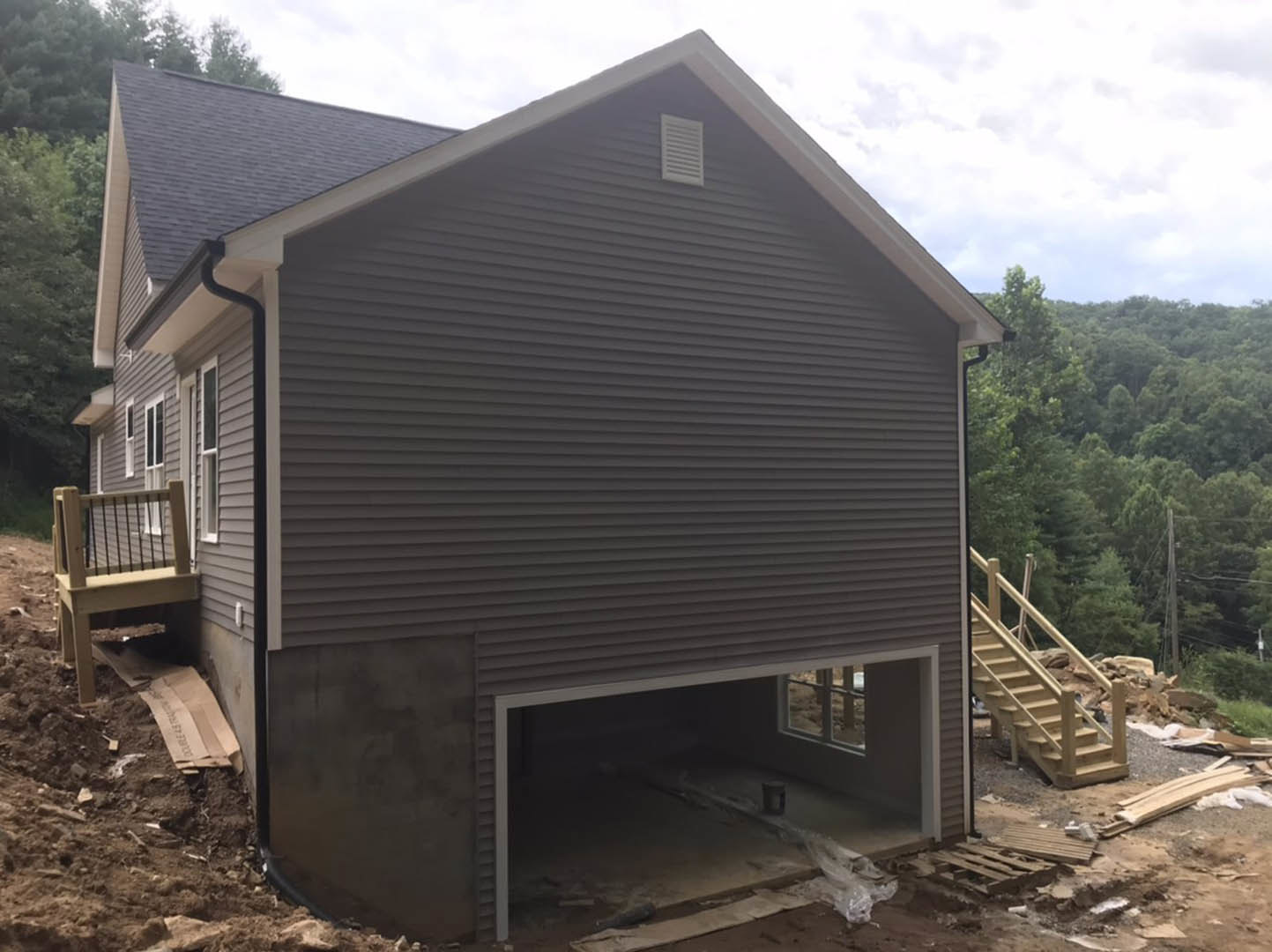 Partially built wooden staircase and deck attached to a house, surrounded by trees, with exposed framing and white vent visible on exterior wall.