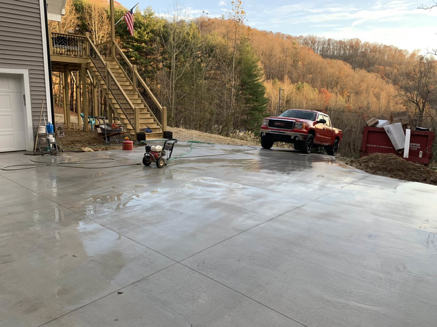 Concrete driveway with a red truck parked beside a red dumpster, American flag on a deck, two-story house with white siding, green lawn, and mature trees in the background