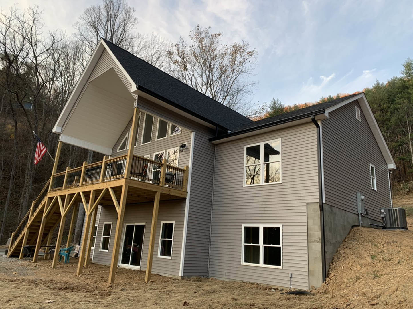 Two-story house with wood siding and white-framed windows, elevated deck with blue chairs, grassy hill and trees in background, American flag near entrance