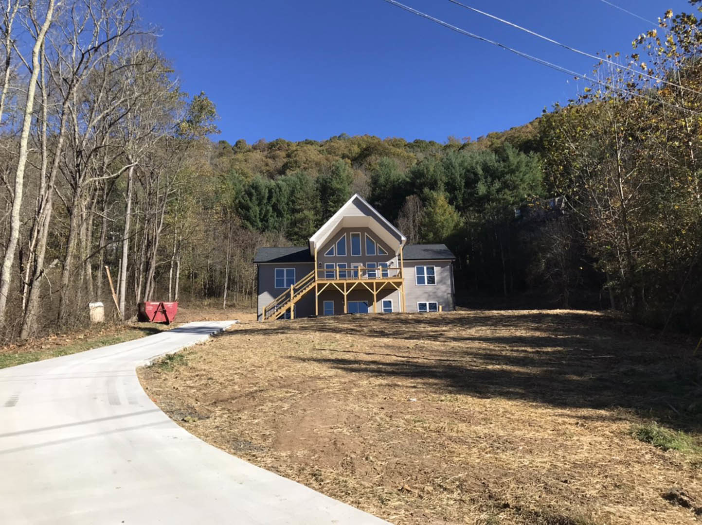 Wide front porch with white railings on a cottage-style house, dirt road leading to the entrance, grassy yard with a red sign, mature trees and blue sky in the background