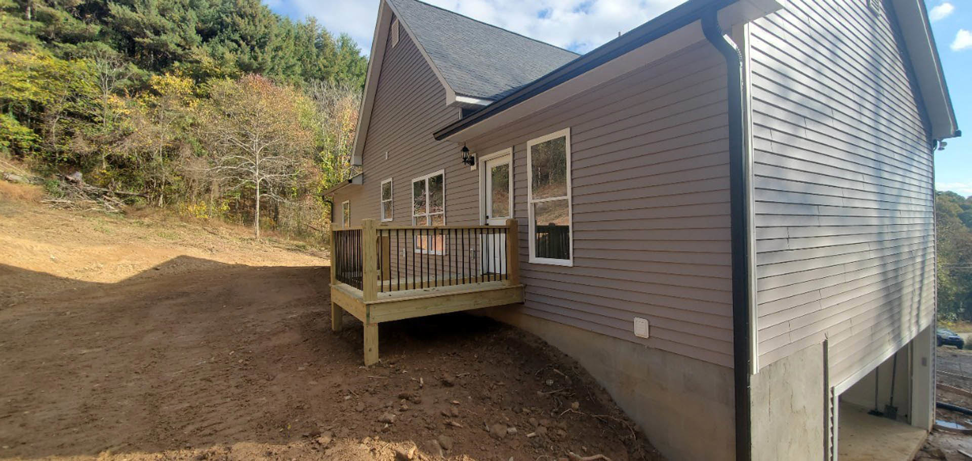 Two-story house with gray siding, wooden deck featuring black metal railing, white-framed windows, covered porch, dirt path leading to entrance, mature trees and blue sky in