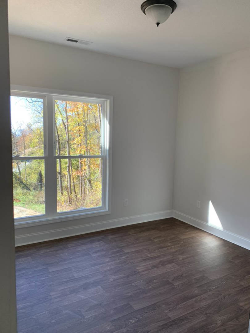Sunlit room with wide window overlooking trees, smooth wood flooring, white plaster walls, and ceiling fan with modern light fixture