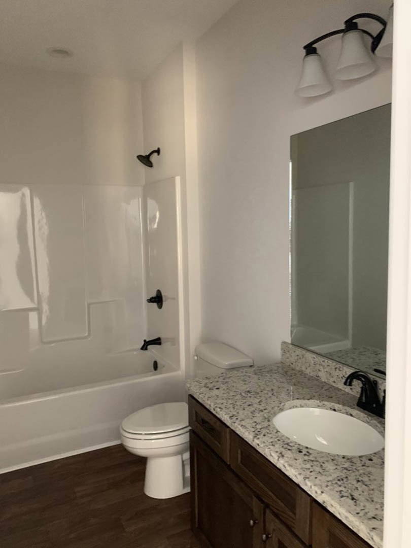 White oval sink set in a stone countertop beside a white toilet and bathtub, light fixture with two white lamps mounted above, neutral tile flooring and walls