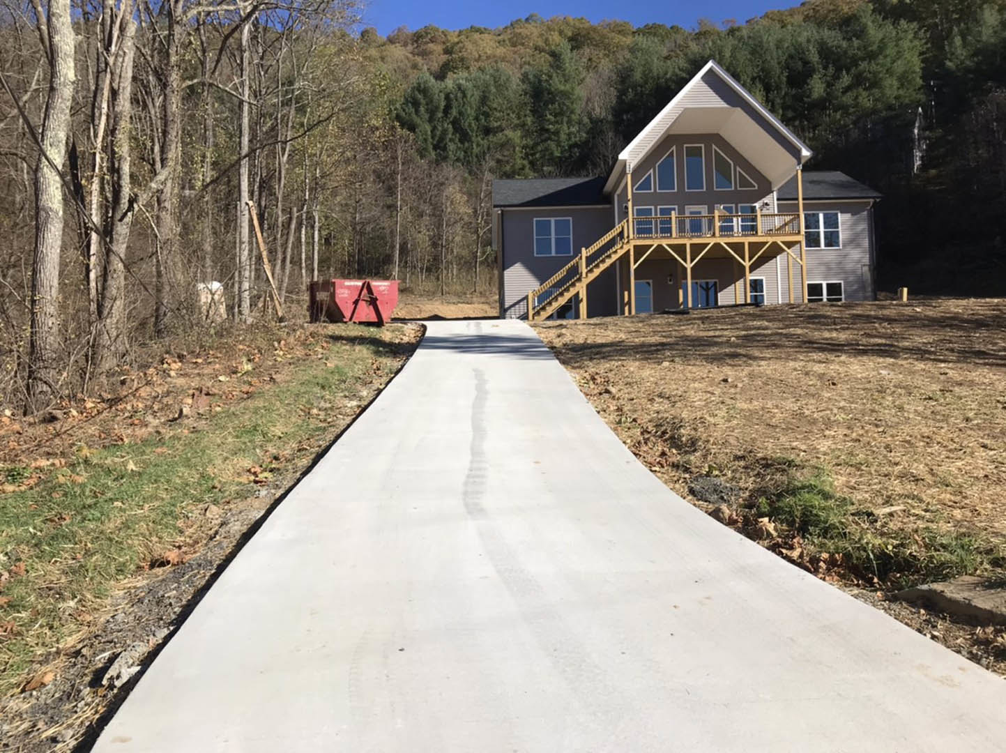 Modern house with wooden deck, white walkway leading to entrance, grassy lawn, mature trees, large red container with triangular design near forested area, black-framed window
