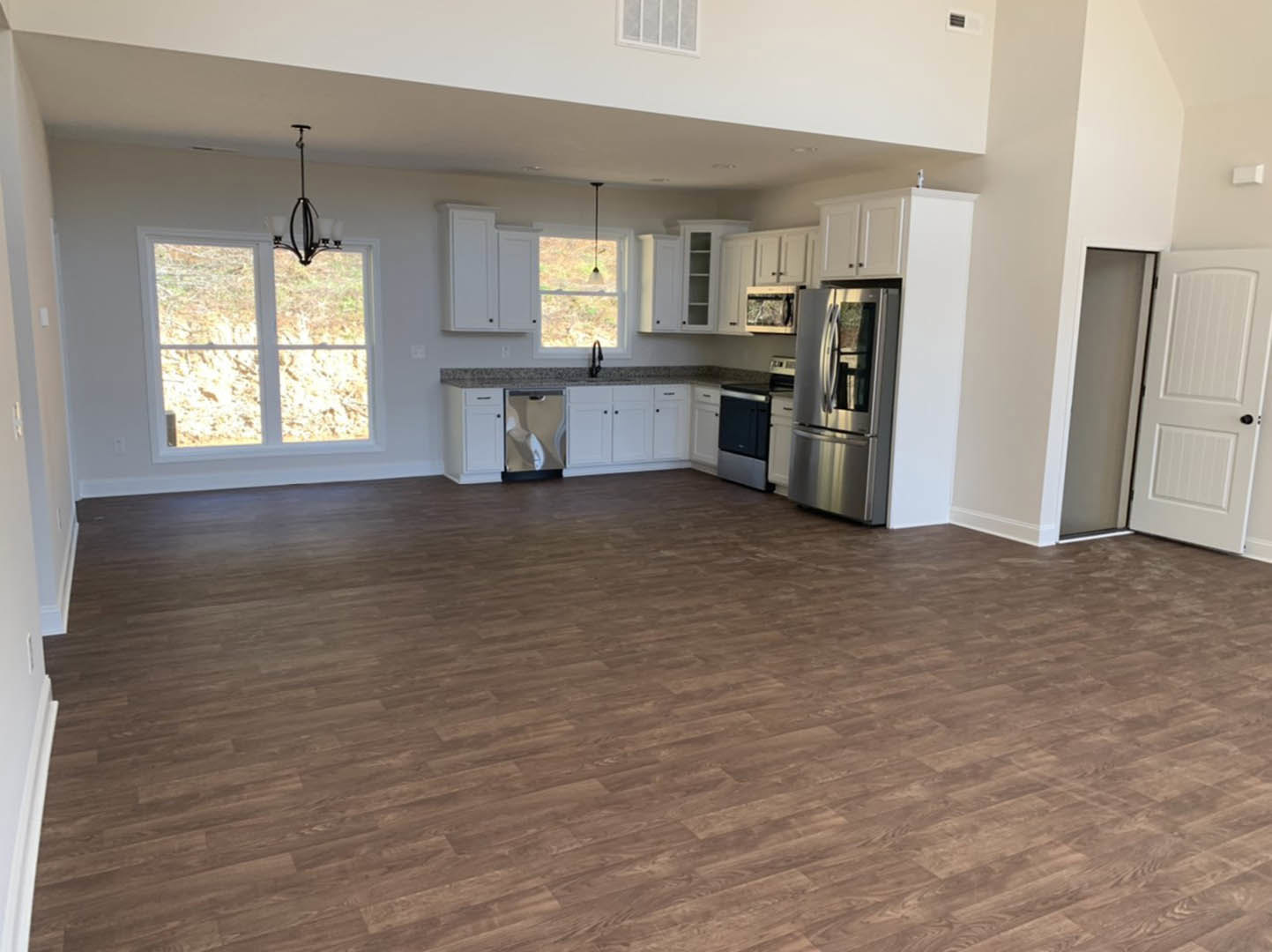 Spacious kitchen and living area featuring wood flooring, white cabinetry, stainless steel appliances, black hardware on a white door, large window with modern light fixture