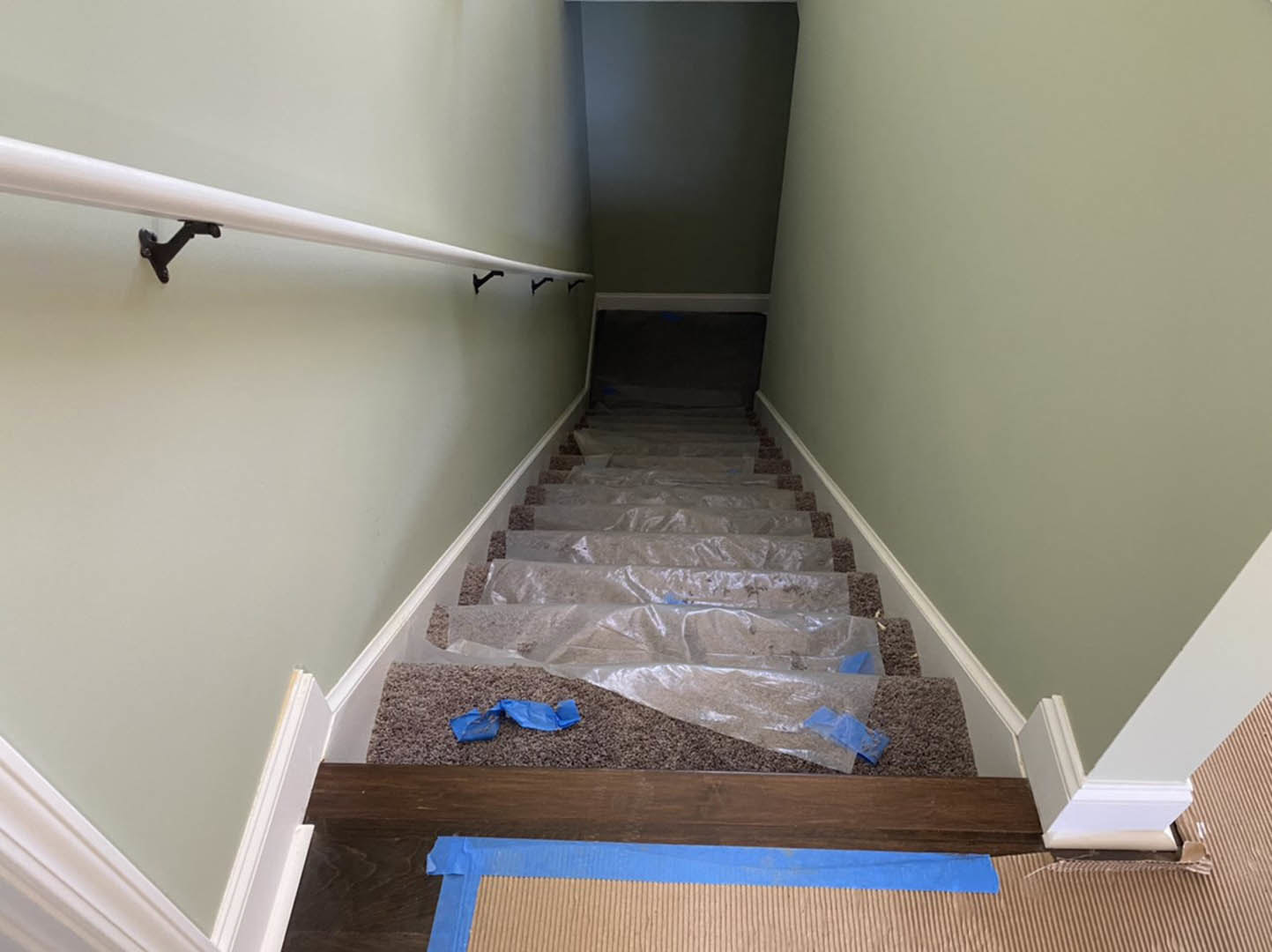 Wooden staircase covered in clear plastic wrap, black metal handrail, white plaster walls, blue and brown striped flooring, white tube mounted on wall, blue object resting on