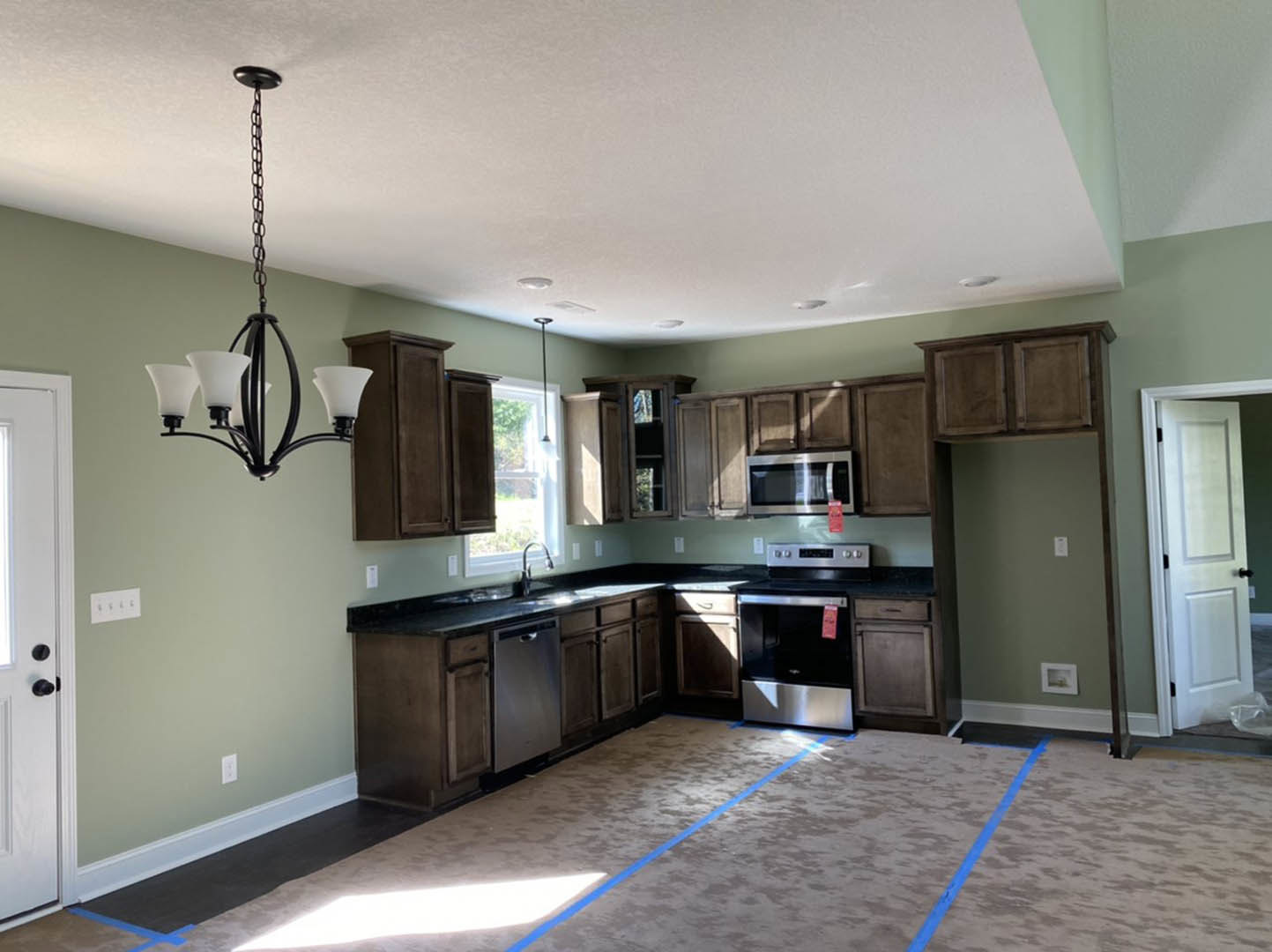 Kitchen with dark wood cabinets, white walls, black switches, stainless steel oven and microwave, modern chandelier, white door with black knobs, light countertops, and sink.