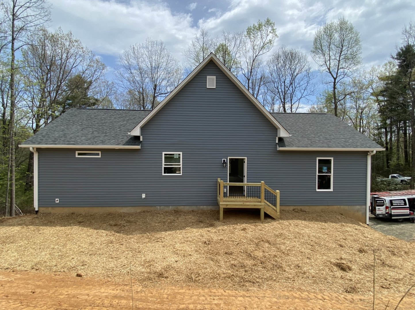 Two-story house with wood deck and porch, wooden stairs leading up, white-framed windows, surrounded by grassy yard and trees under cloudy sky