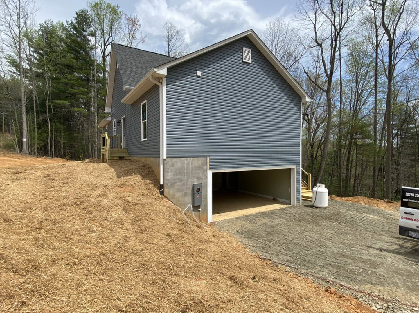 Two-story house with attached garage, light siding, dark roof, mature trees in background, gravel driveway, white propane tank on dirt area