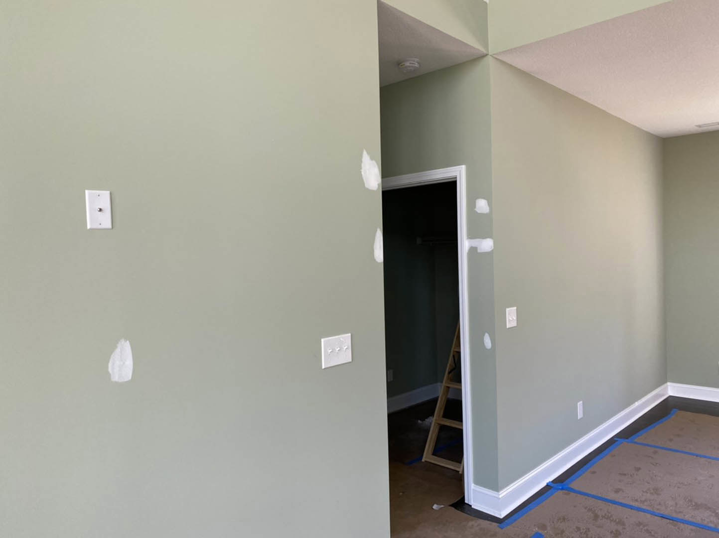 Open room with white plaster walls, blue painter’s tape on light-colored floor, two ladders, white wall plate with round silver fixture, and white triple light switch.