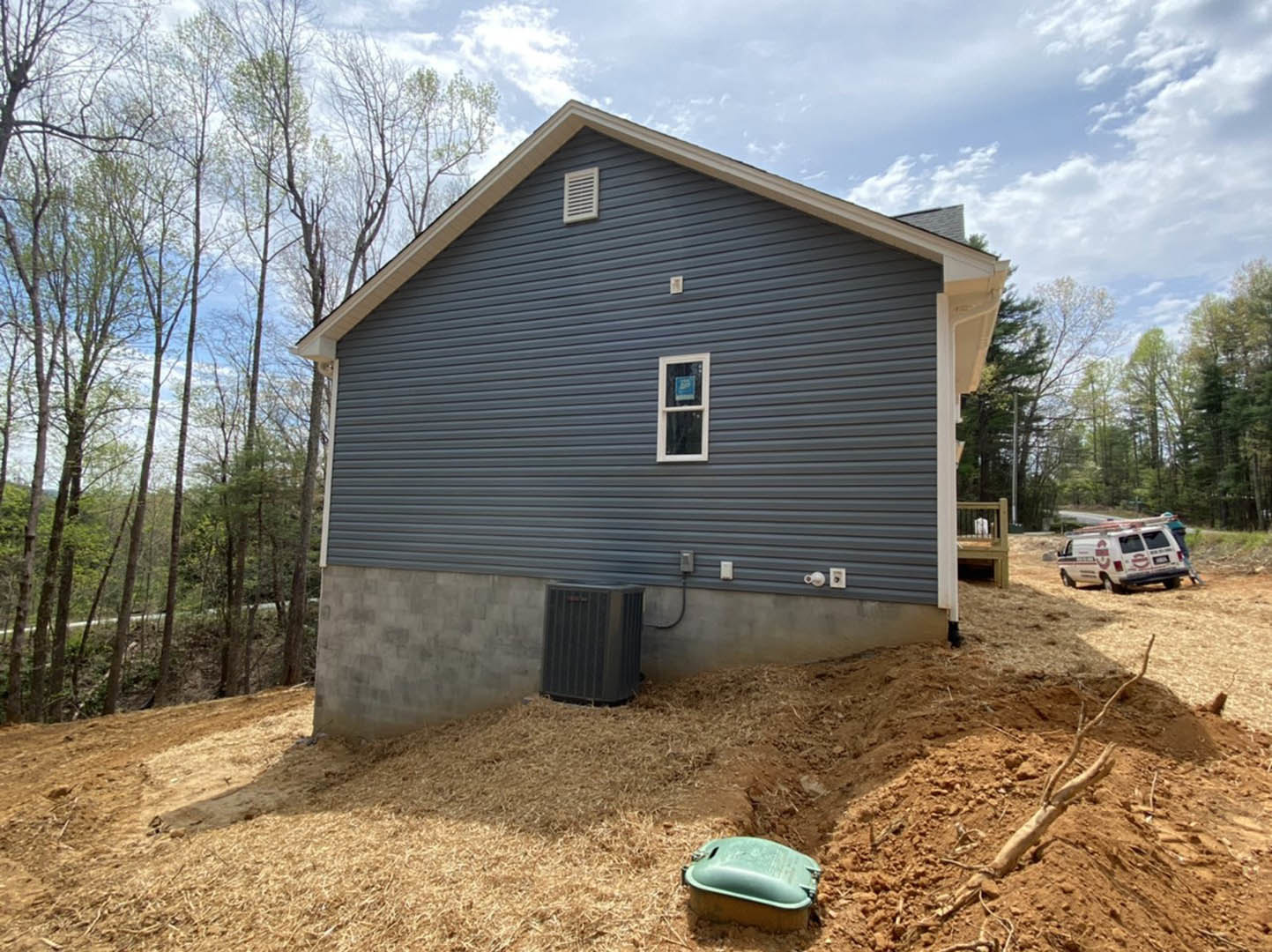 Modern house with large windows, surrounded by dirt hill and mature trees, green container and black metal grill in foreground, white van with red and white logos parked nearby
