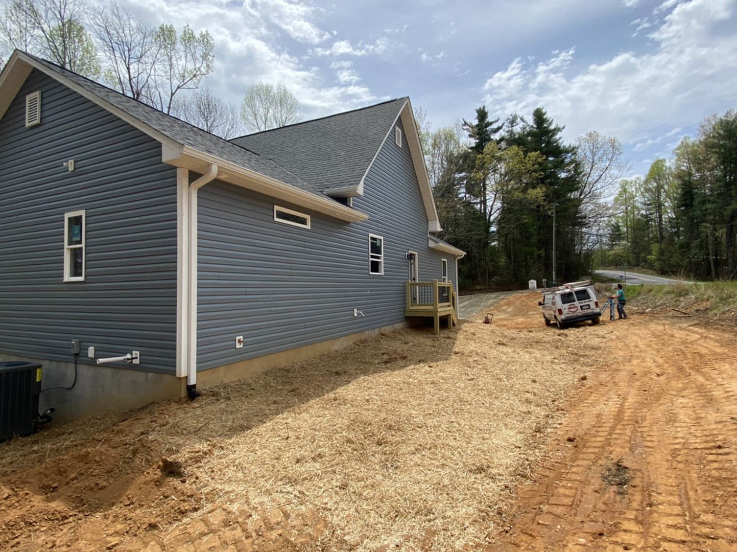 Blue siding house with wooden porch railing, elevated deck, grassy yard, white van with red and white logos parked behind, person walking on ladder near exterior.