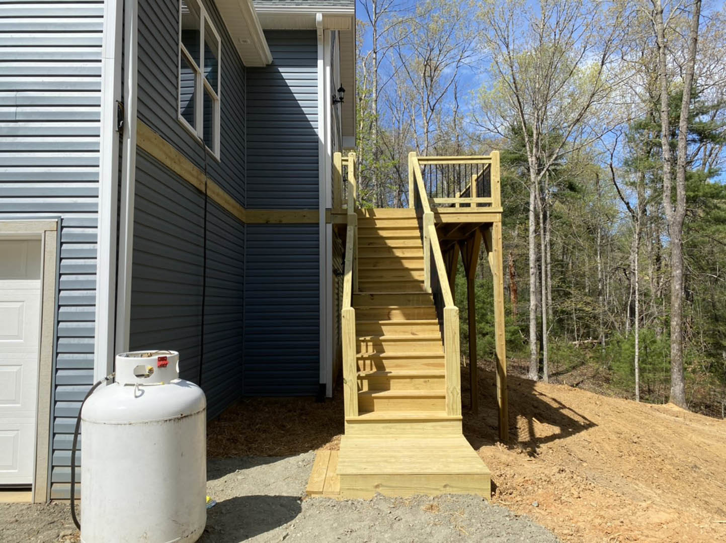 Wooden deck stairs leading to a white door with metal handle, surrounded by trees; propane tank and waste container positioned near the house exterior.