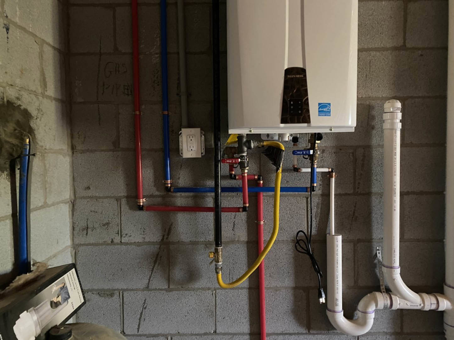 White tankless water heater mounted on wall with exposed copper pipes, electrical outlet, and nearby utility boxes in unfinished interior space