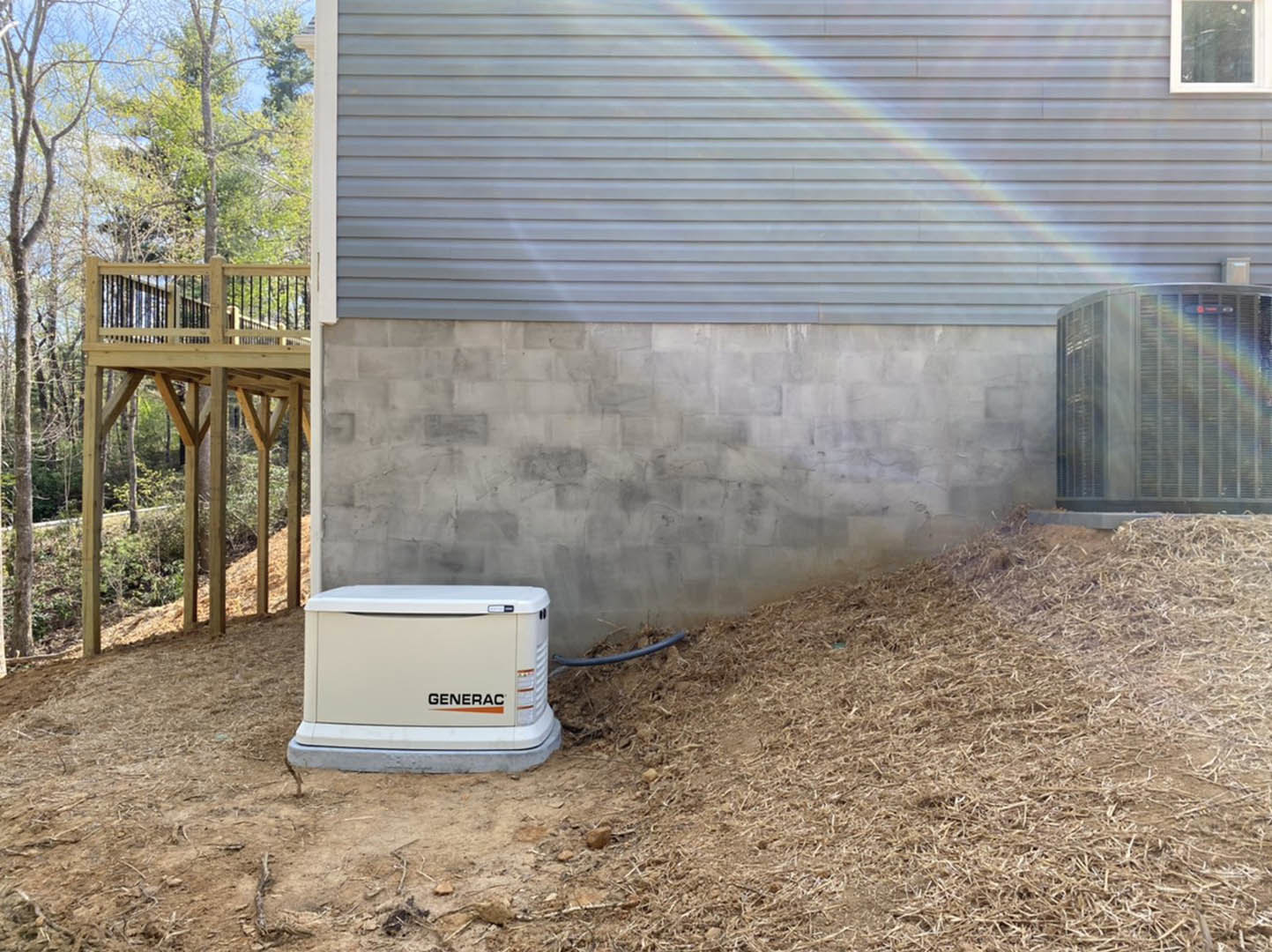 White generator with logo and red indicator light installed beside wooden deck, rainbow mural on exterior wall, white-framed window, trees in background.