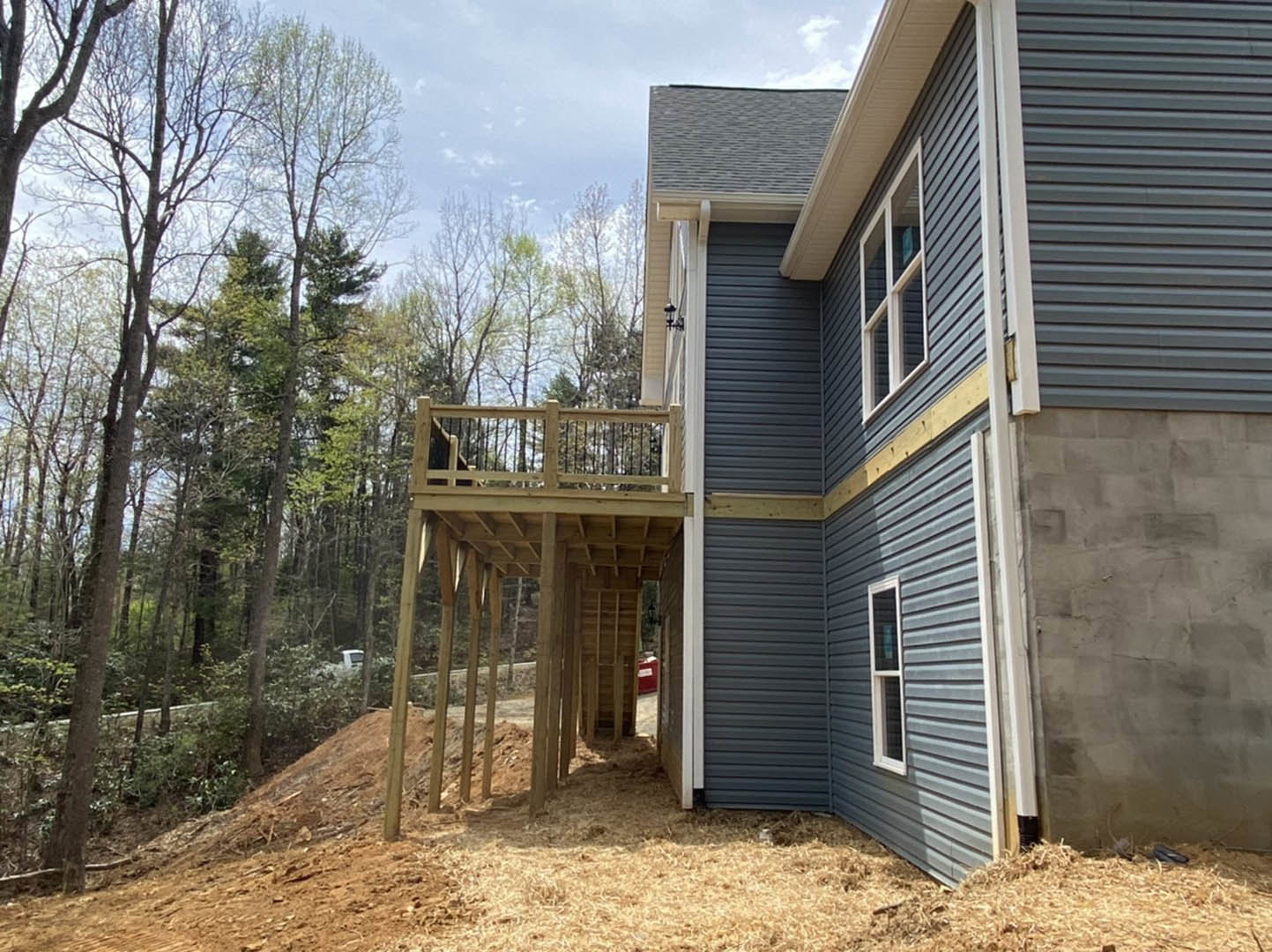 Wood-framed house under construction featuring a partially built deck and balcony, large windows, and surrounded by tall trees