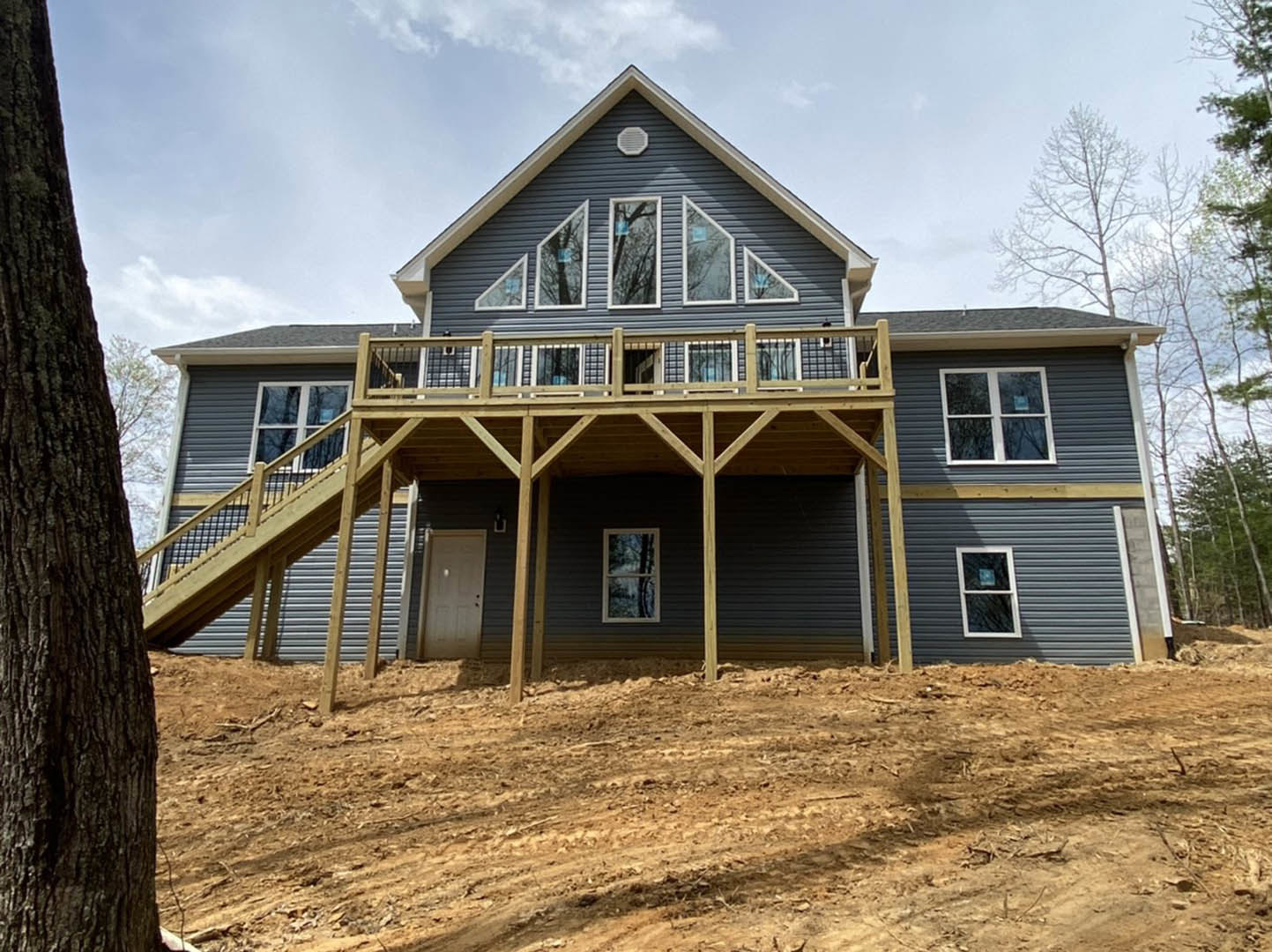 Two-story home with light siding, elevated wooden deck, staircase leading to backyard, large windows with white frames, mature trees and dirt field in background