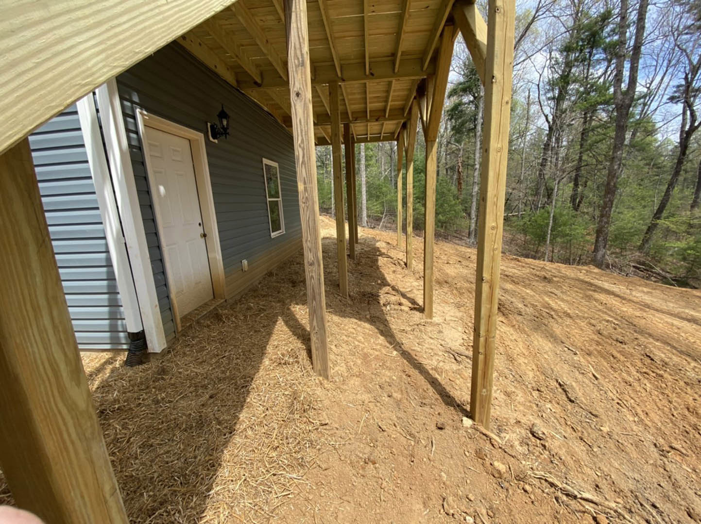 Covered porch with grey siding, white front door, adjacent window, wooden support posts, dirt ground, and surrounding trees