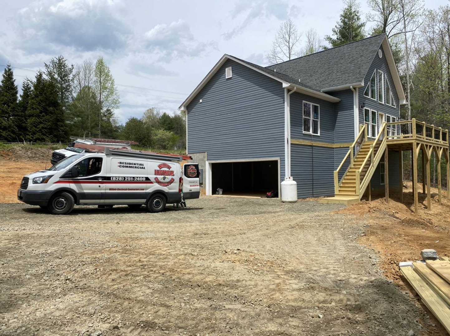 White truck parked on driveway in front of two-story house with attached garage, exterior stairs leading to elevated deck, white siding, red accents, and surrounding green lawn.