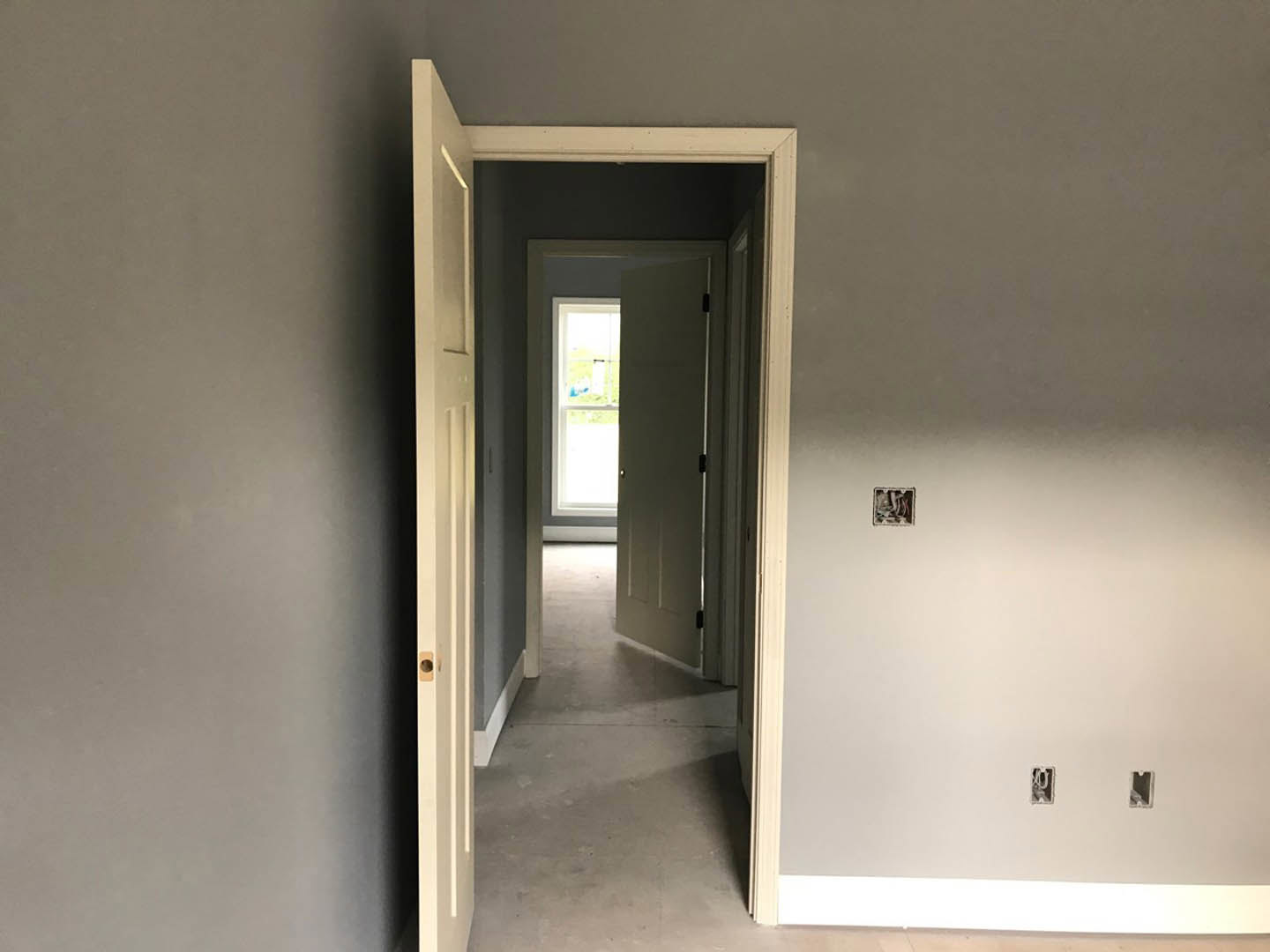 Hallway with light-colored plaster walls, open door revealing window with white frame, hardwood flooring, and partial view of a person's leg