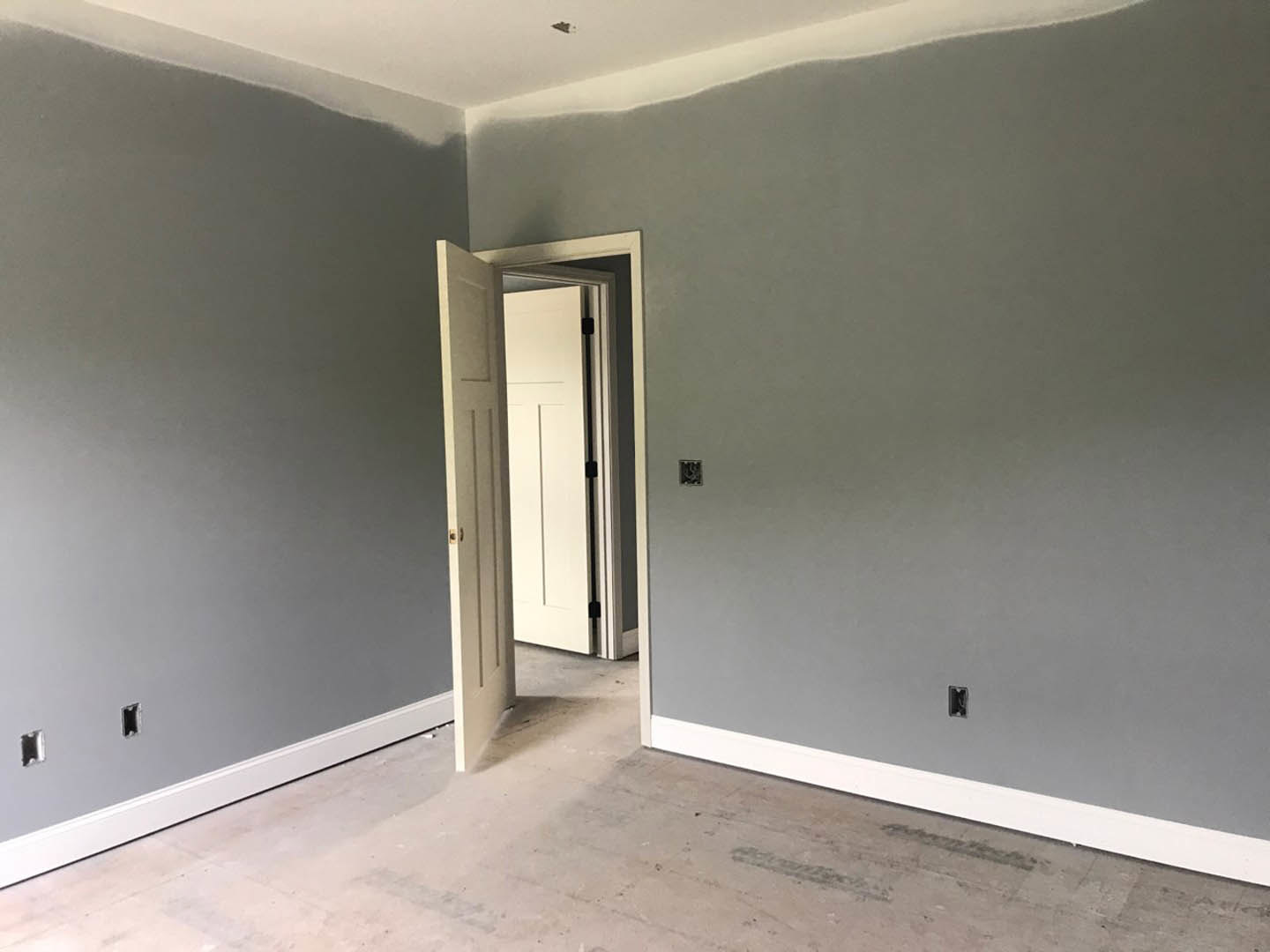 White paneled door with black handle opening into a room with light wood flooring, white walls, and subtle grey accents.