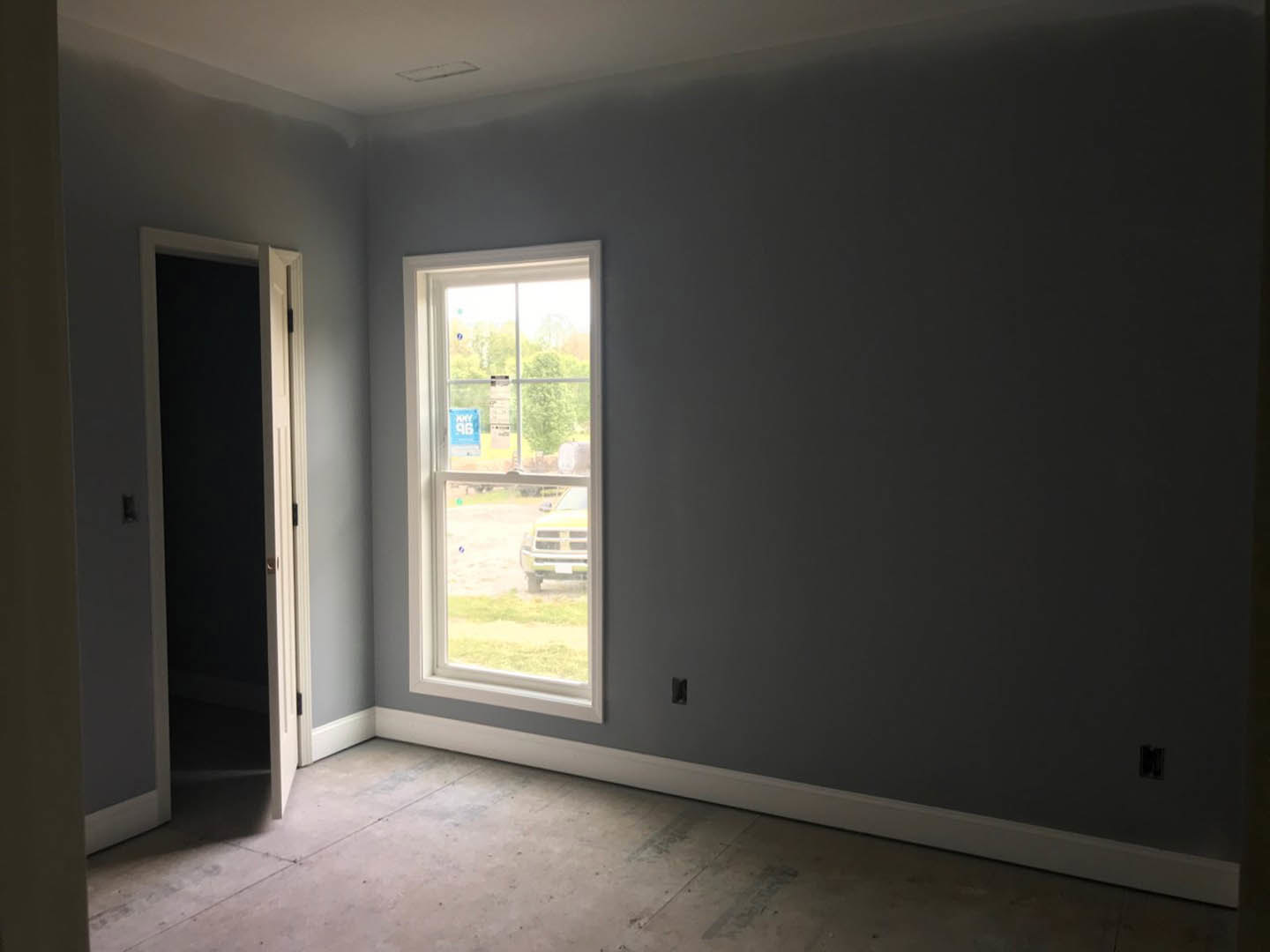 Neutral-toned room featuring plaster walls, wood flooring, large window with a sign, and a white door; yellow truck visible outside through the window
