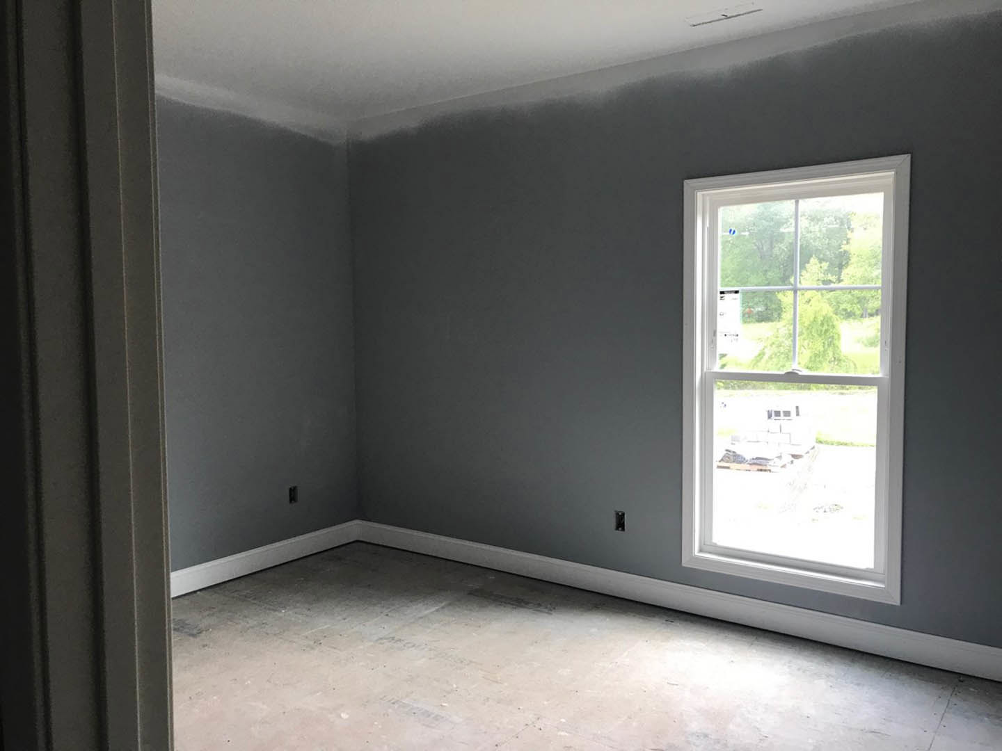 Sunlit room with smooth plaster walls, large window overlooking trees and grass, polished concrete floor, and minimalist ceiling fixture.