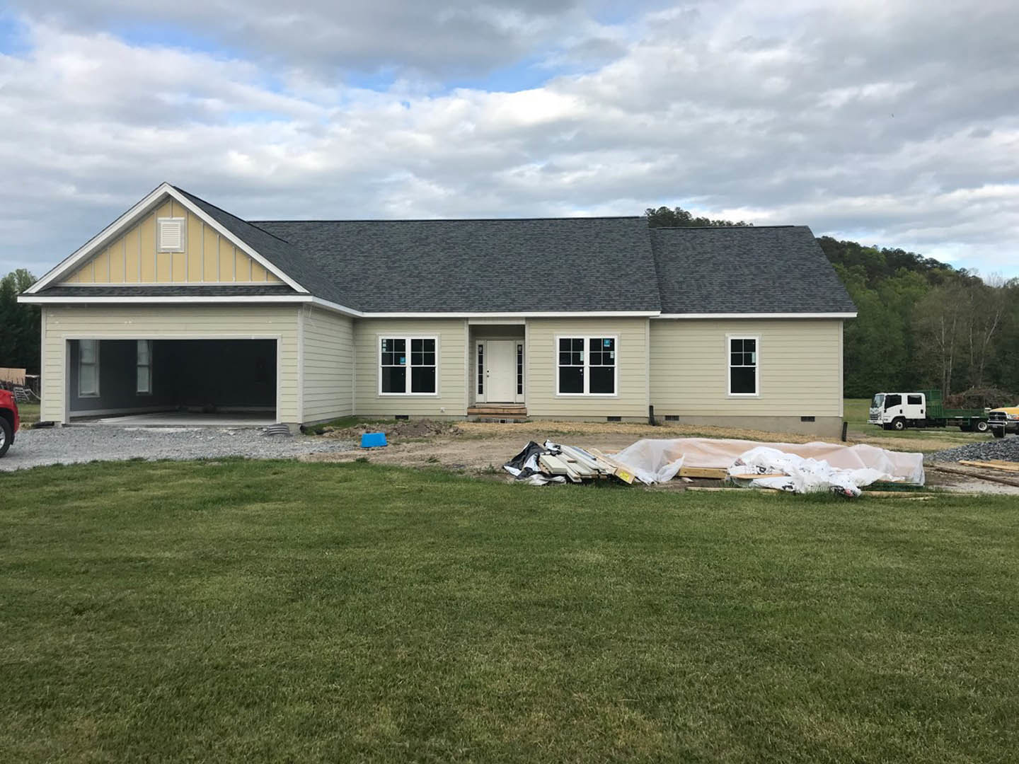 Two-story house with white door and window, green grass yard, pile of wood and plastic debris, cloudy sky in background, white truck with green container parked nearby
