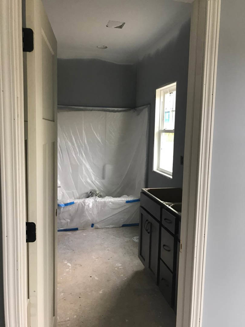 Bathroom under renovation with white plastic sheet draped over surfaces, concrete floor marked with blue tape, white cabinet, window displaying blue sign, and unfinished plaster