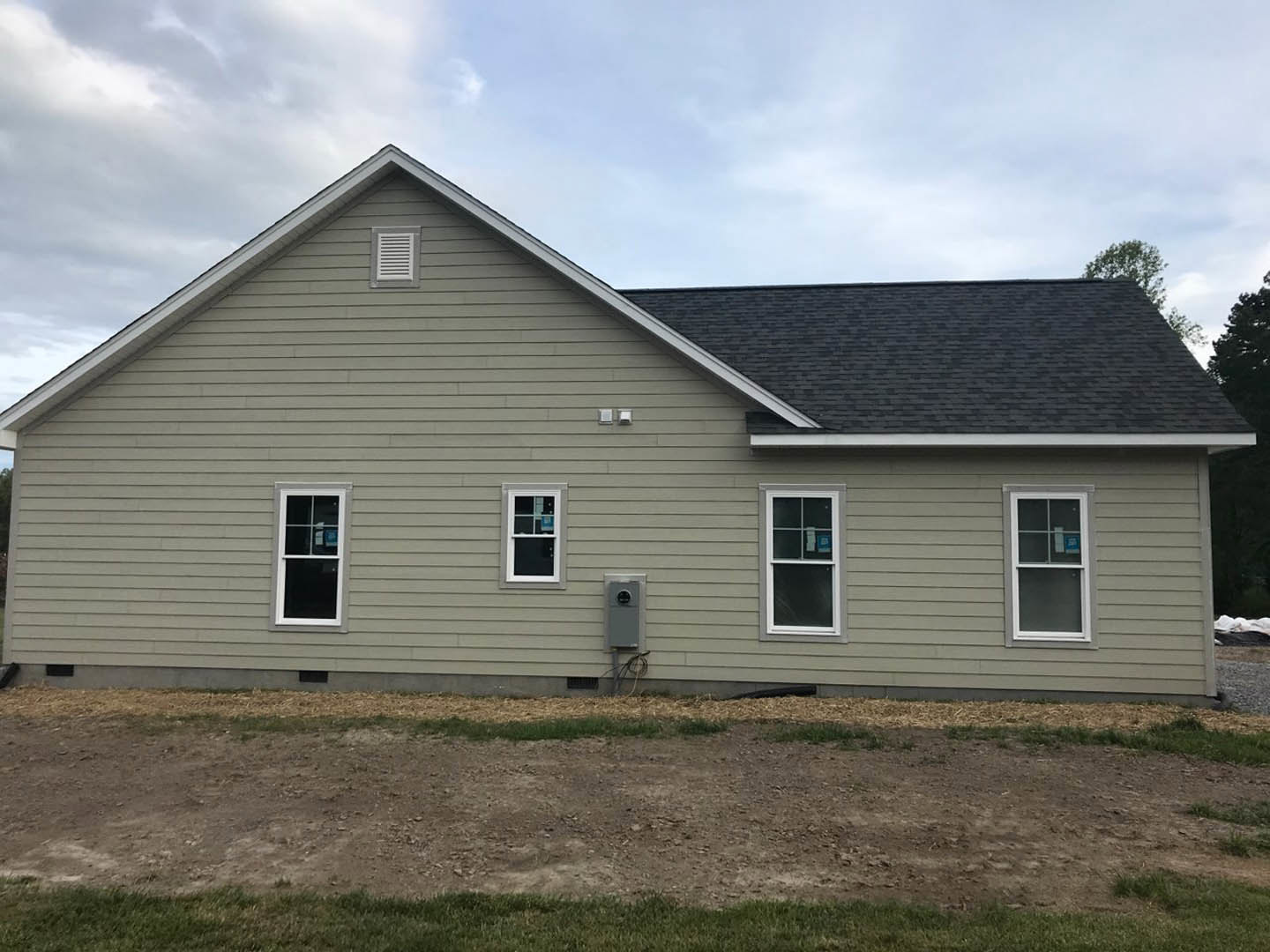 Two-story house with grey roof, white-framed windows, and light siding, set behind a dirt path bordered by patchy grass, under a cloudy sky.