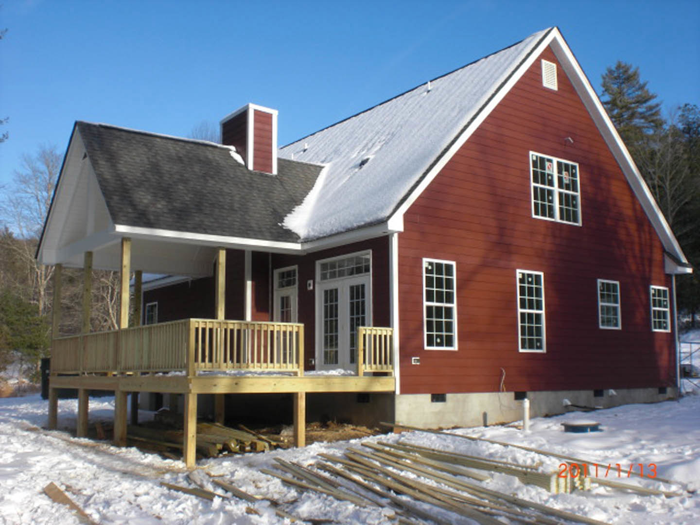 Two-story house with wood deck and railing, snow covering ground and roof, multiple square windows, bare trees in winter landscape