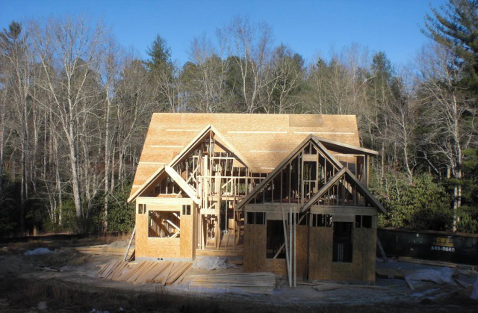 Wood-framed house under construction surrounded by tall trees, exposed beams and window opening visible beneath clear blue sky
