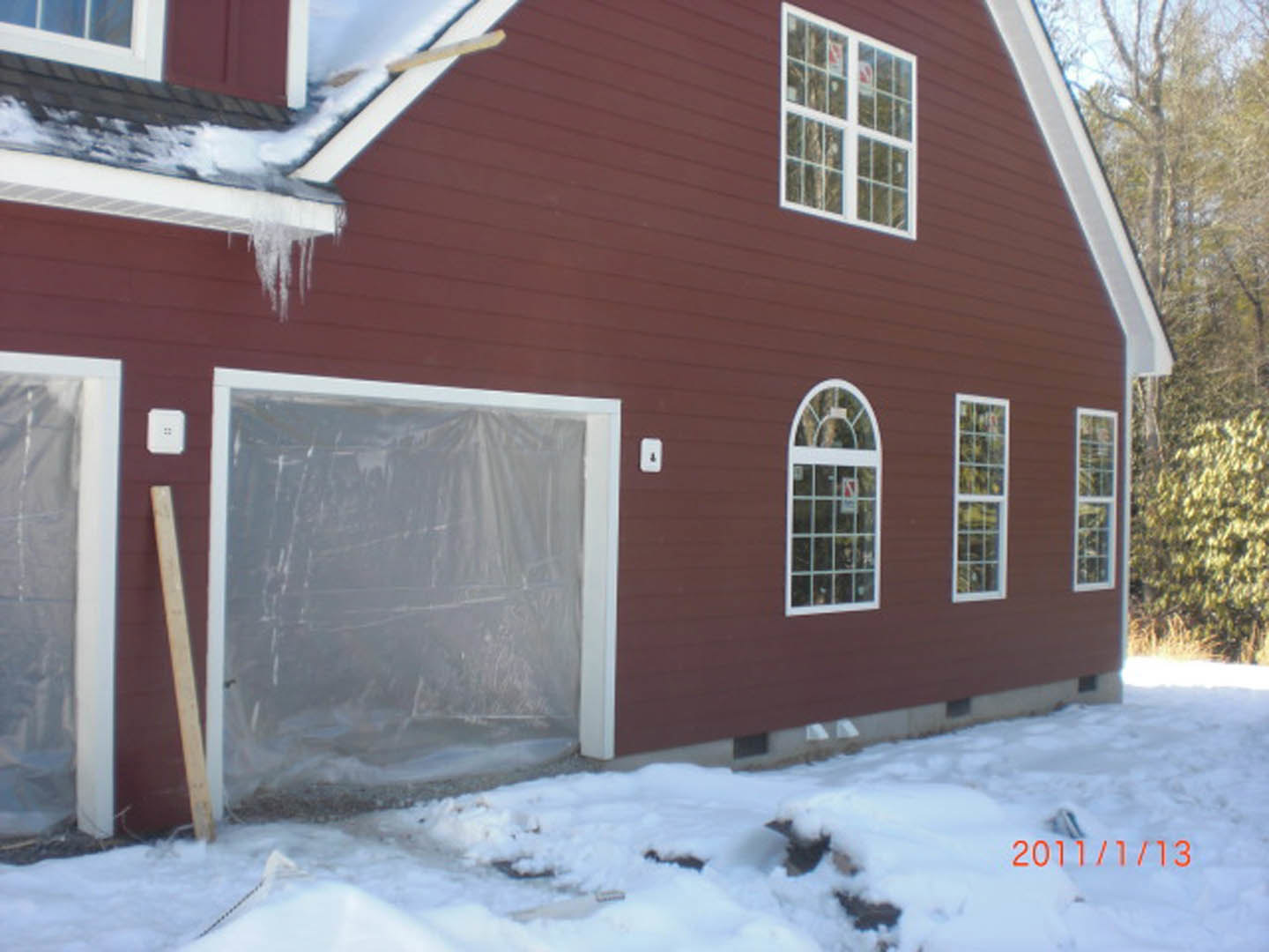 Two-story home with white siding, attached garage door, snow-covered ground, rectangular windows, and bare trees in the background