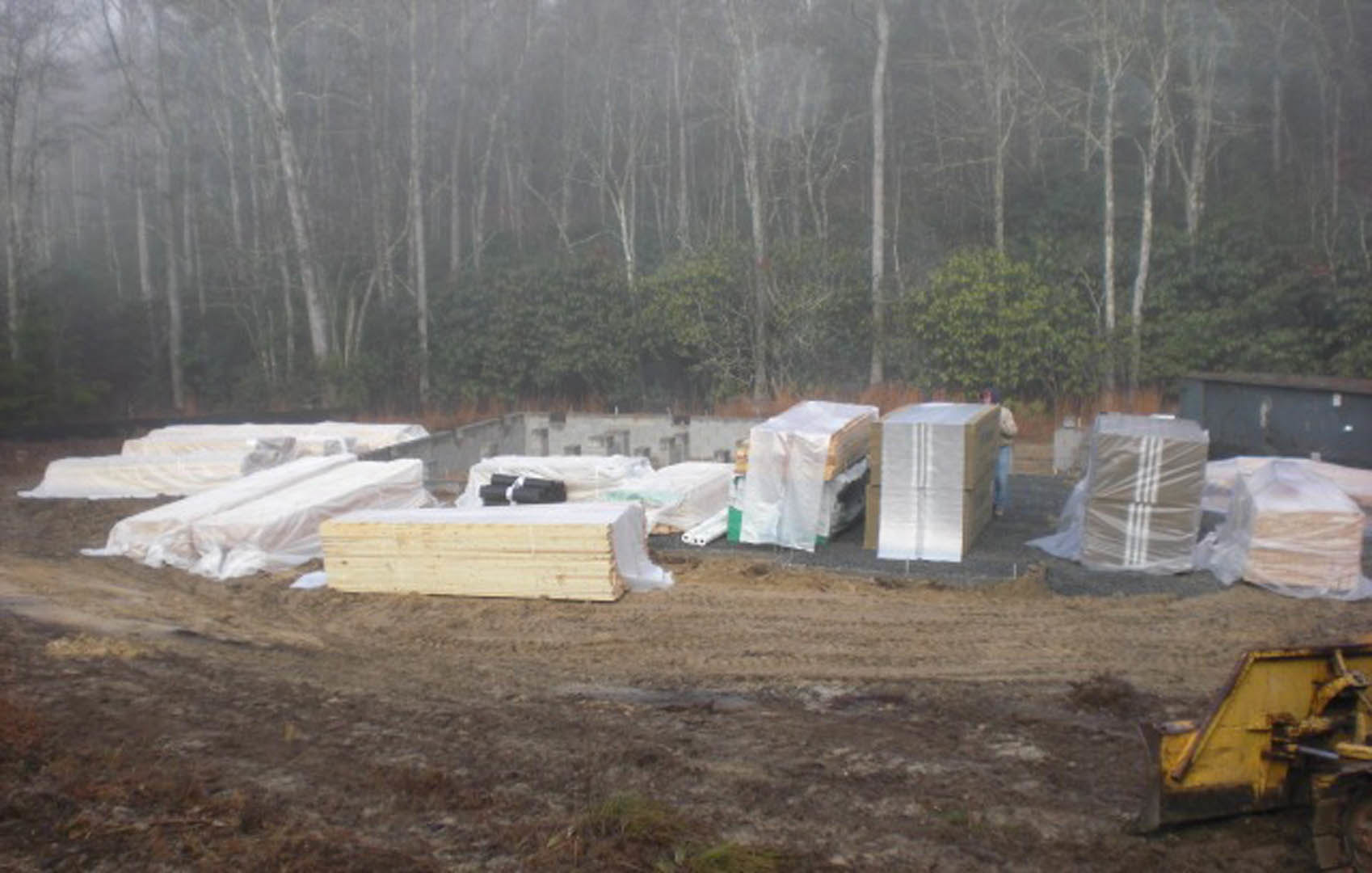 Wood pallets and white plastic sheeting covering foundation at residential construction site, surrounded by grass and trees