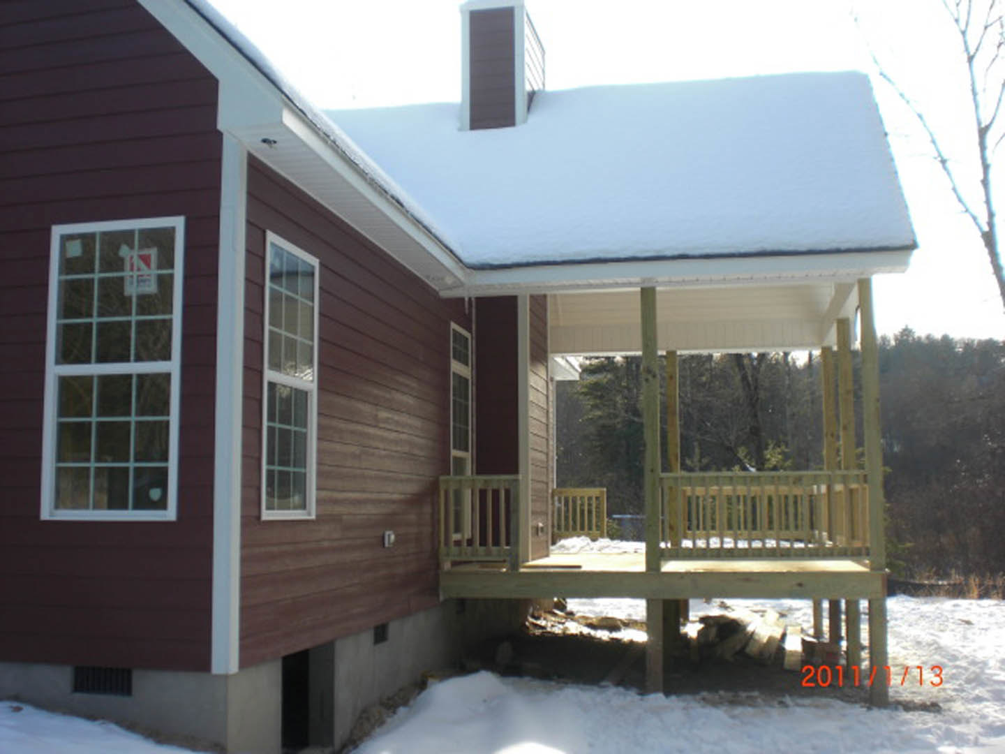 Two-story home with white siding, multi-pane windows, and a wooden deck overlooking a snow-covered yard; roof and fence also dusted with snow, bare trees visible in the background.