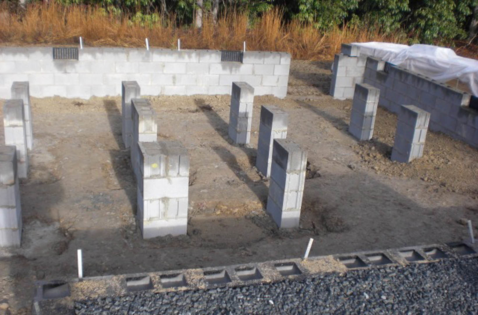 Concrete foundation with upright columns, surrounded by grass and a white brick wall, close-up of drainage system visible, white tarp covering part of block wall