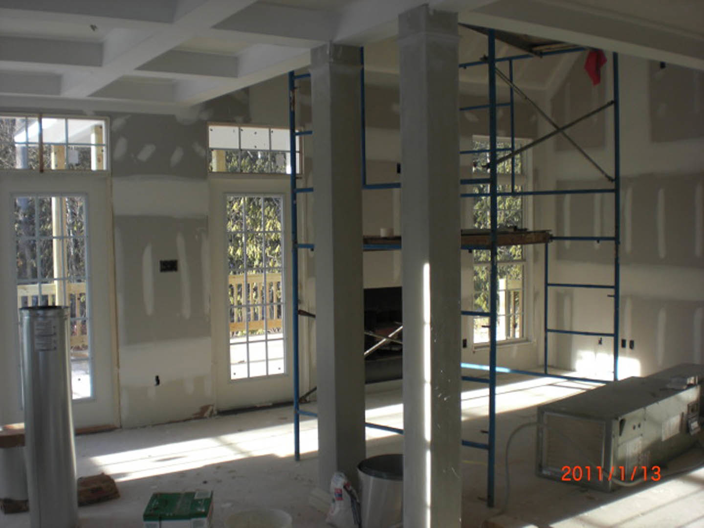 Spacious room featuring a floor-to-ceiling window framed in aluminum, white walls, exposed ceiling beams, and sheer white curtains allowing natural daylight.