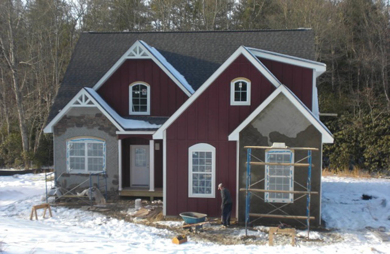 Snow-covered modern cottage with large windows, wood siding, and a man standing near the front door; leafless trees and scaffolding visible in the yard.
