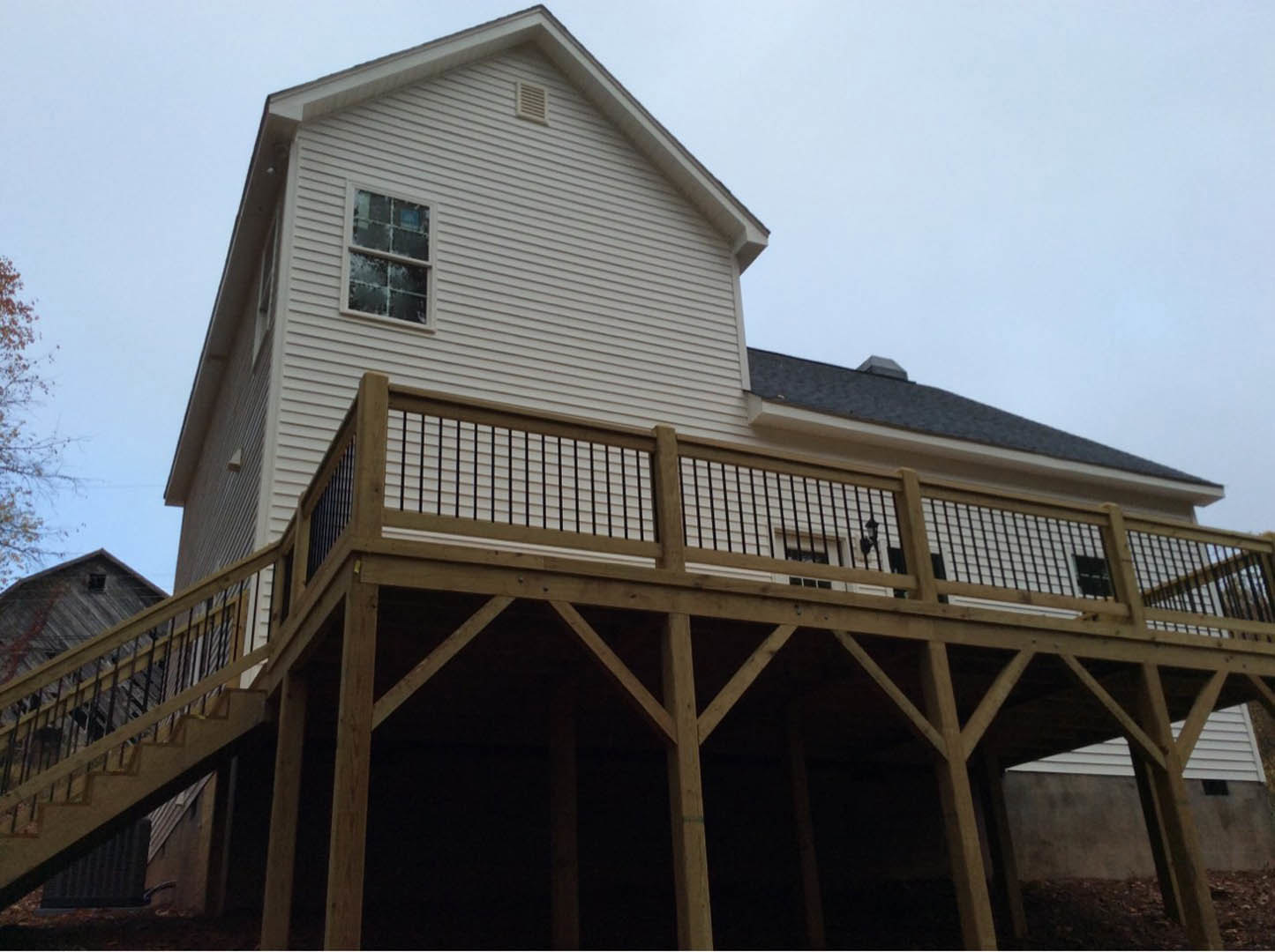Wooden deck with white railing attached to a house, white-framed window, gray siding, tree branches and blue sky in background, shingled roof visible above
