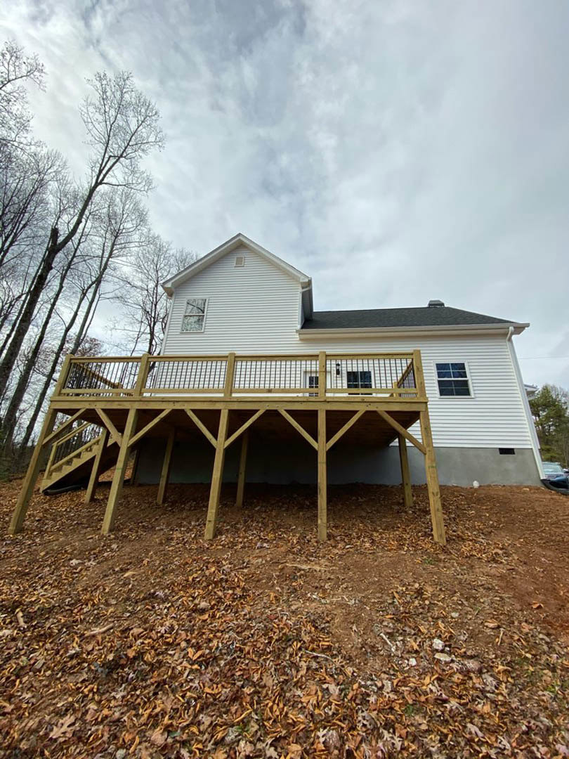 Two-story cottage with multi-pane windows, wooden deck featuring metal railing, scattered leaves on ground, surrounded by tall trees under partly cloudy sky