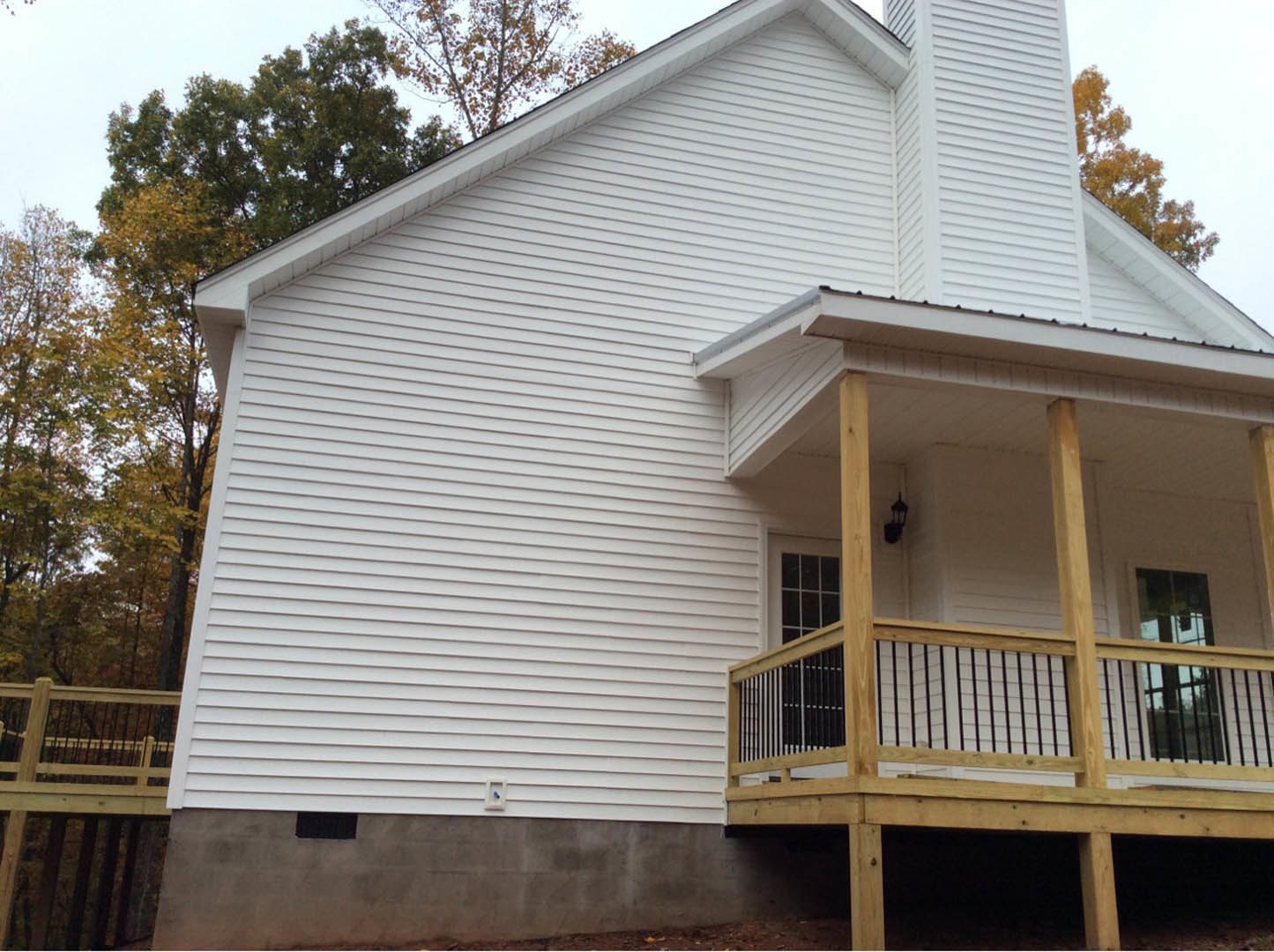 White siding home with covered porch, wooden deck and railing, large window framed by wood beam, leafy tree in foreground