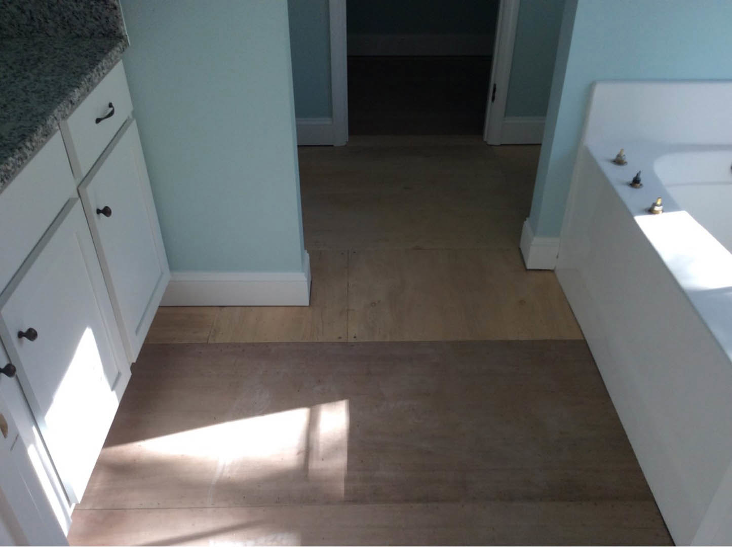 Modern kitchen with granite countertops, stainless steel sink beneath a large window, white cabinetry, tile backsplash, and wood flooring illuminated by natural light.