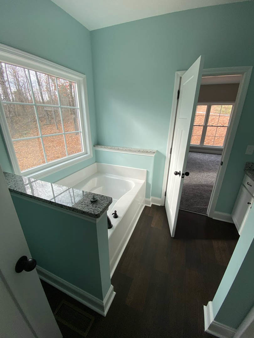 Bathroom with freestanding white bathtub beneath large window overlooking forest, light gray tile flooring, chrome faucet, white walls, and open door leading to adjacent room