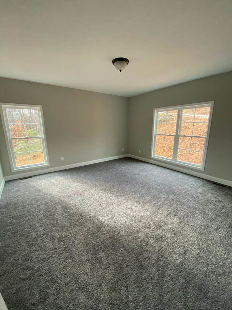 Bright carpeted room with large windows, white plaster walls, ceiling-mounted light fixture, and views of green trees outside