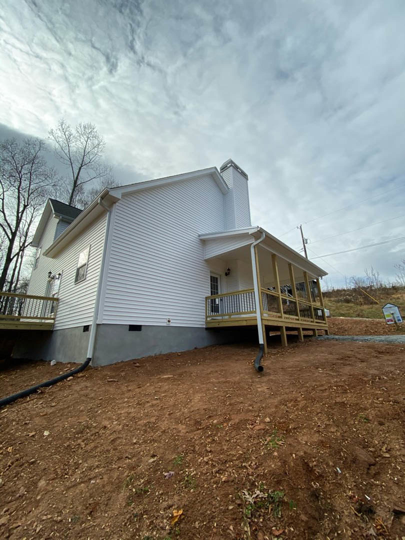 Two-story house with light siding, covered front porch, elevated wooden deck, dirt hill with black pipe, grassy yard, cloudy sky