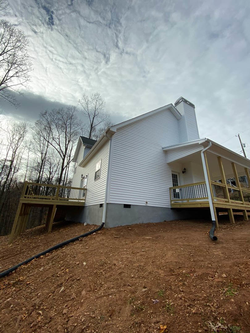 Two-story house with light siding, wooden deck with railing, covered front porch, dirt yard with black pipe, trees and cloudy sky in background