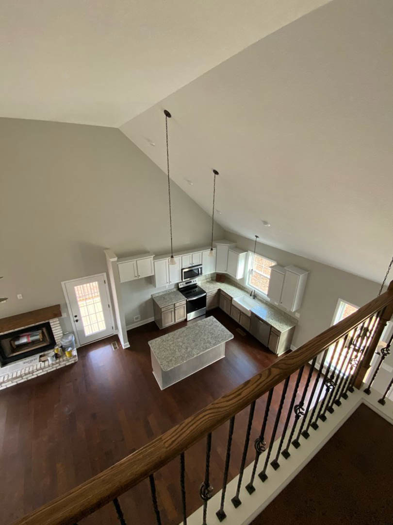 Open kitchen and living room with hardwood floors, marbled rectangular dining table, white cabinetry, stainless steel stove and oven, staircase with wood handrail, and white door