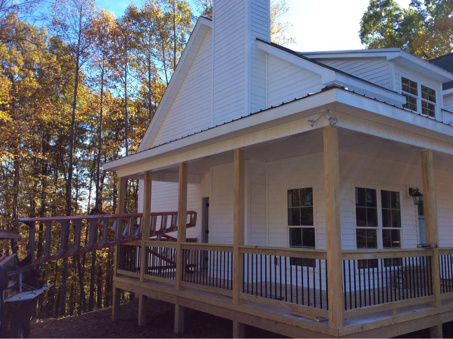 Wood siding home with a covered porch featuring a wooden railing, white-framed window, ladder, and slide; surrounded by trees under a sloped roof.