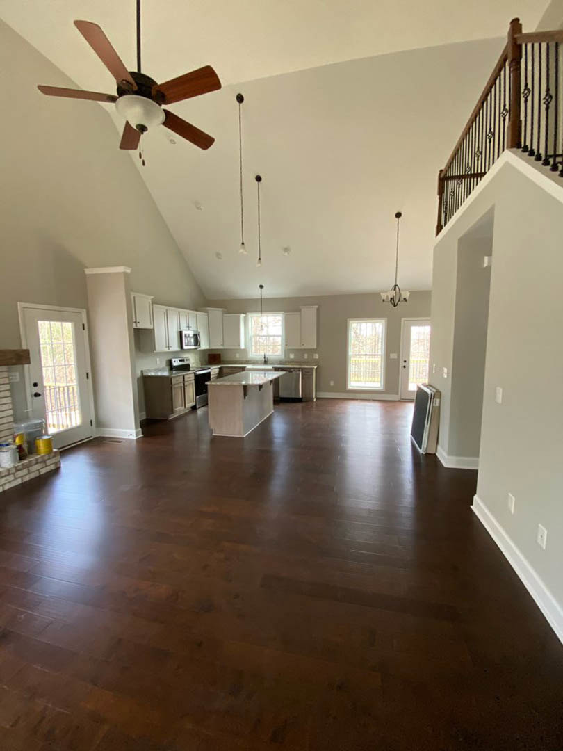 Spacious open floor plan featuring wood flooring, kitchen with large white island and countertop, ceiling fan with light fixture, glass-paneled door, and staircase visible in