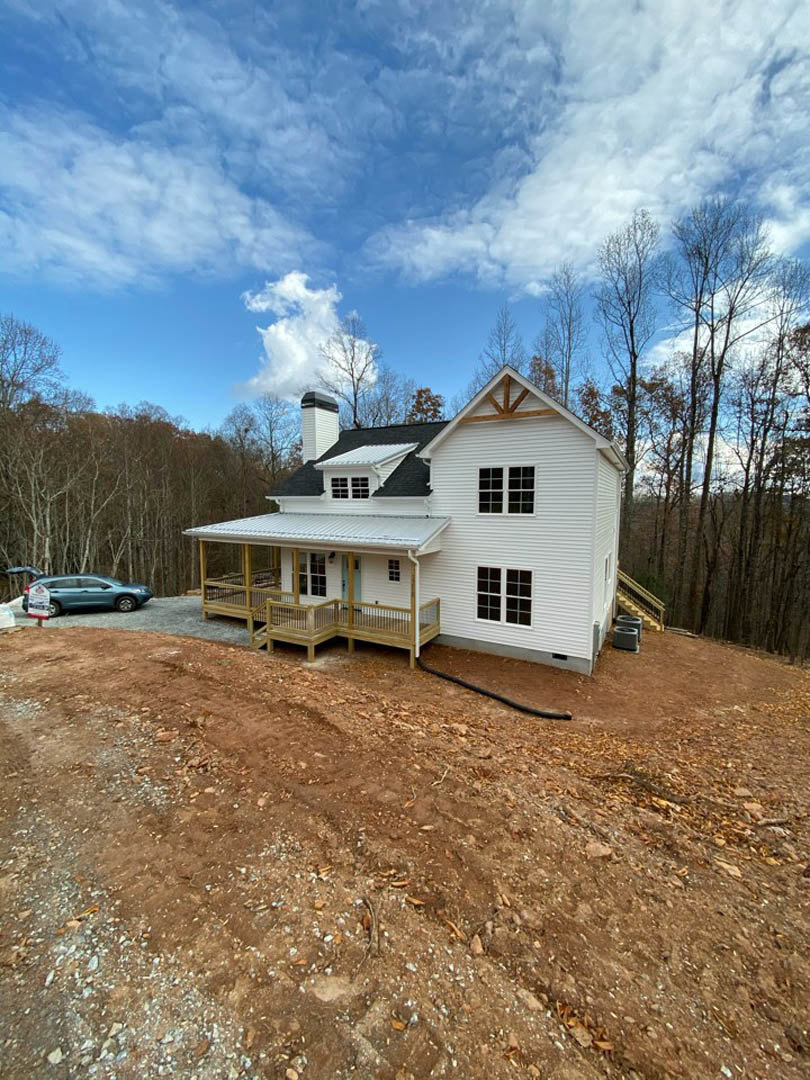 White cottage-style house with porch and railing, chimney, and deck, situated on a grassy hill with mature trees in the background; dirt path and car visible near the road.